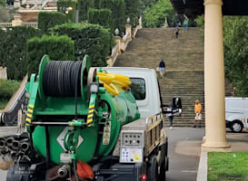 Camión de vacío verde de Tub-Net estacionado frente a unas escaleras de piedra, con carrete de manguera y señales de advertencia en la parte trasera